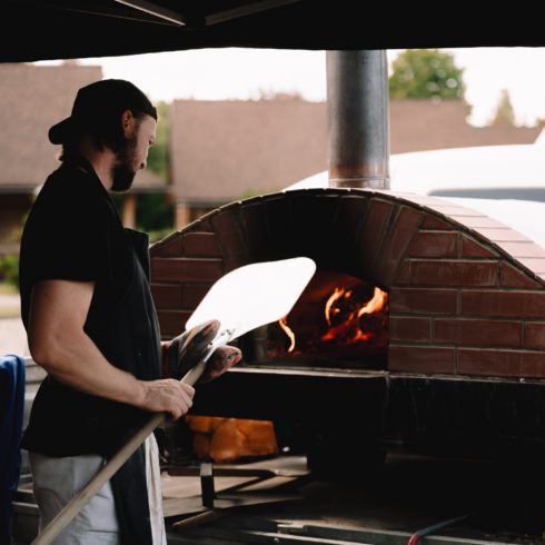 Izbraukuma ēdināšana ar malkas picu krāsni - picu cepējs ar lāpstu rokās